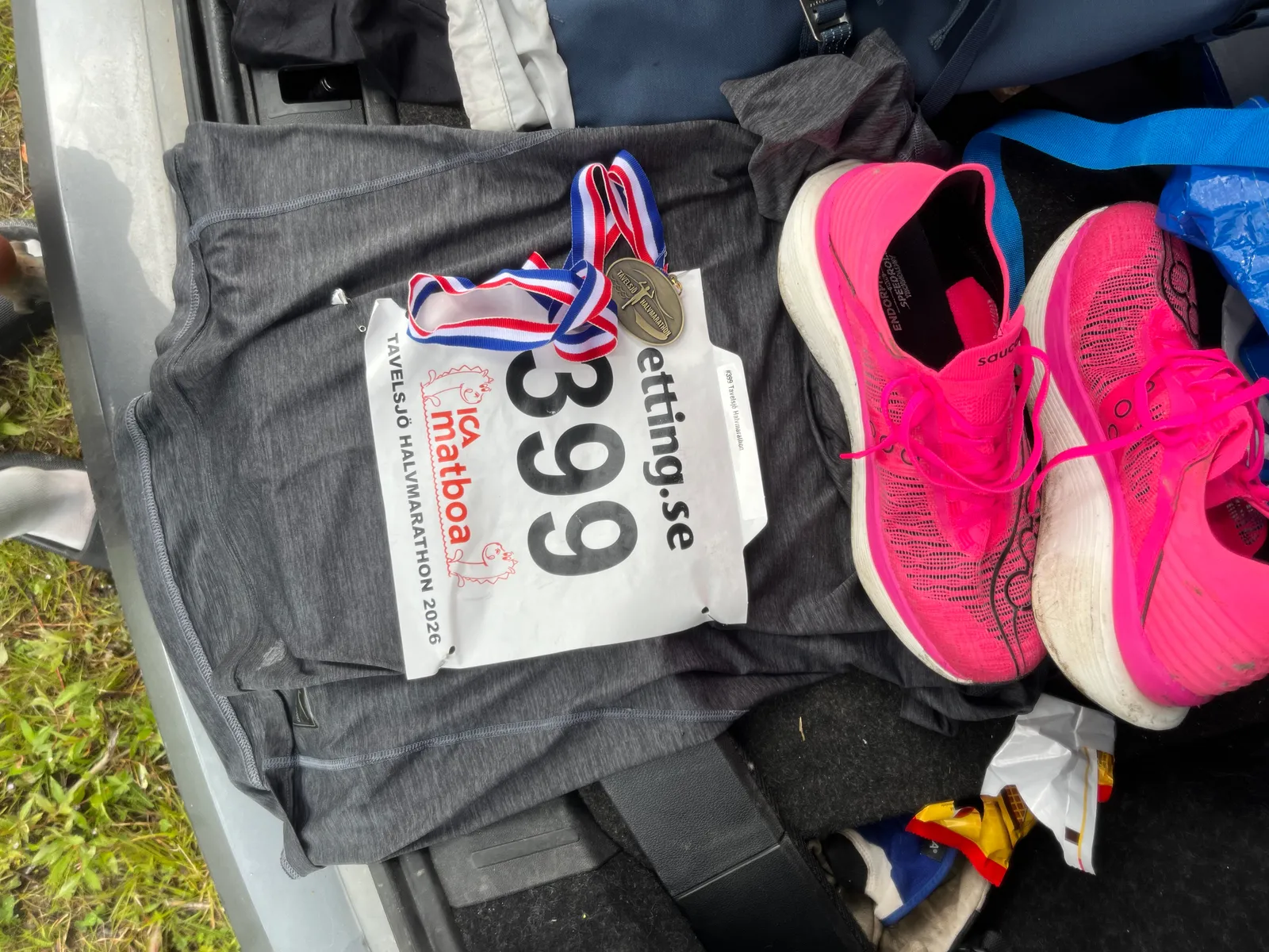 Race bib number 399 for the Tavelsjö Halvmarathon 2026, a finisher's medal and a pair of pink racing shoes laid out in a car boot