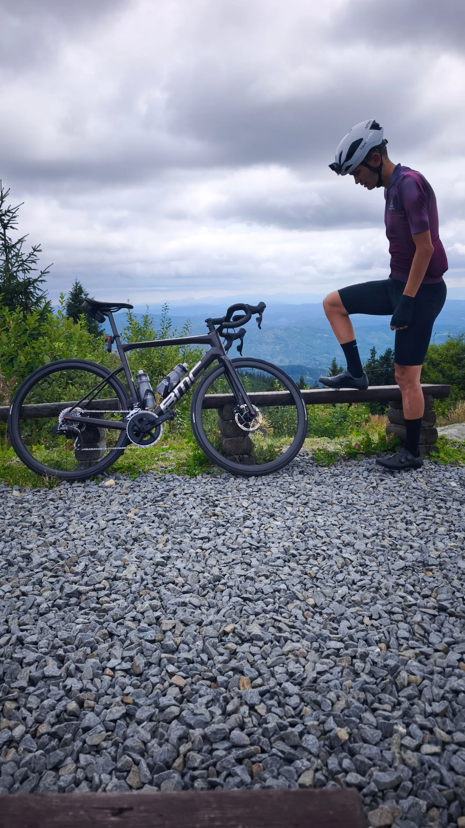Juraj beside his road bike at a summit, with forested ridgelines behind him under heavy cloud