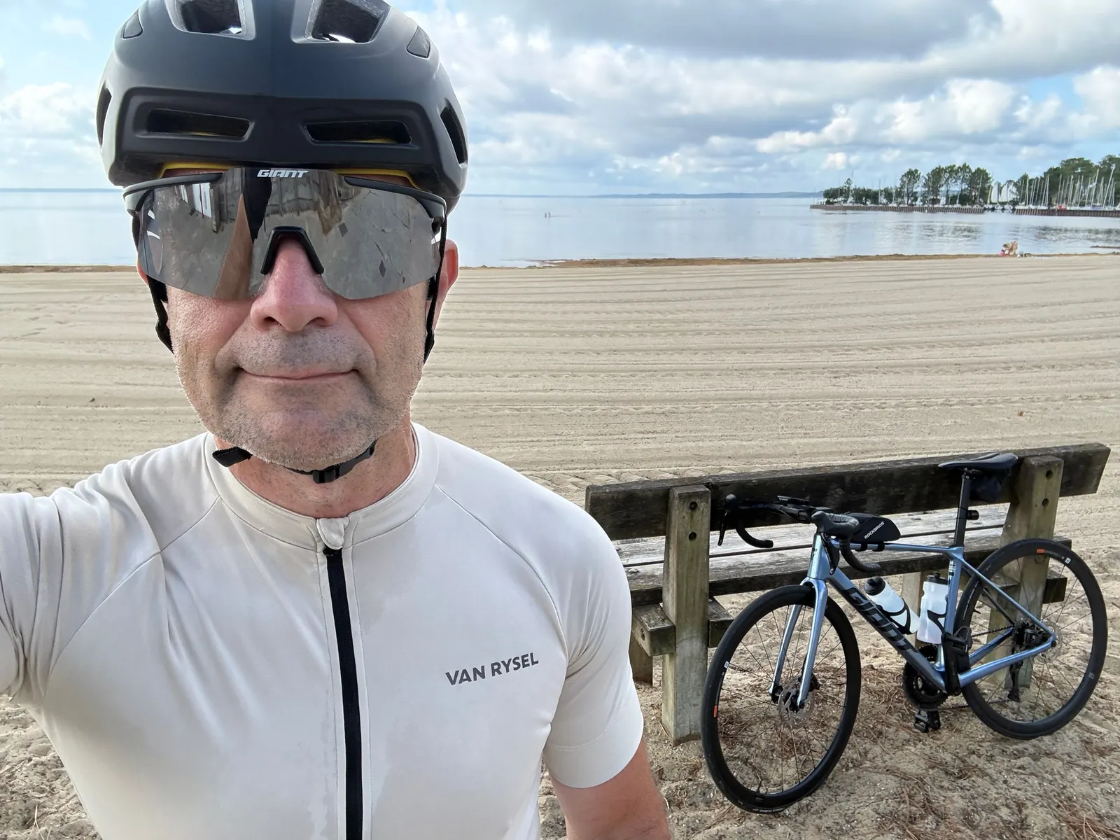 Olivier in a cycling helmet and sunglasses on a sandy beach, with his road bike leaning on a wooden bench behind him