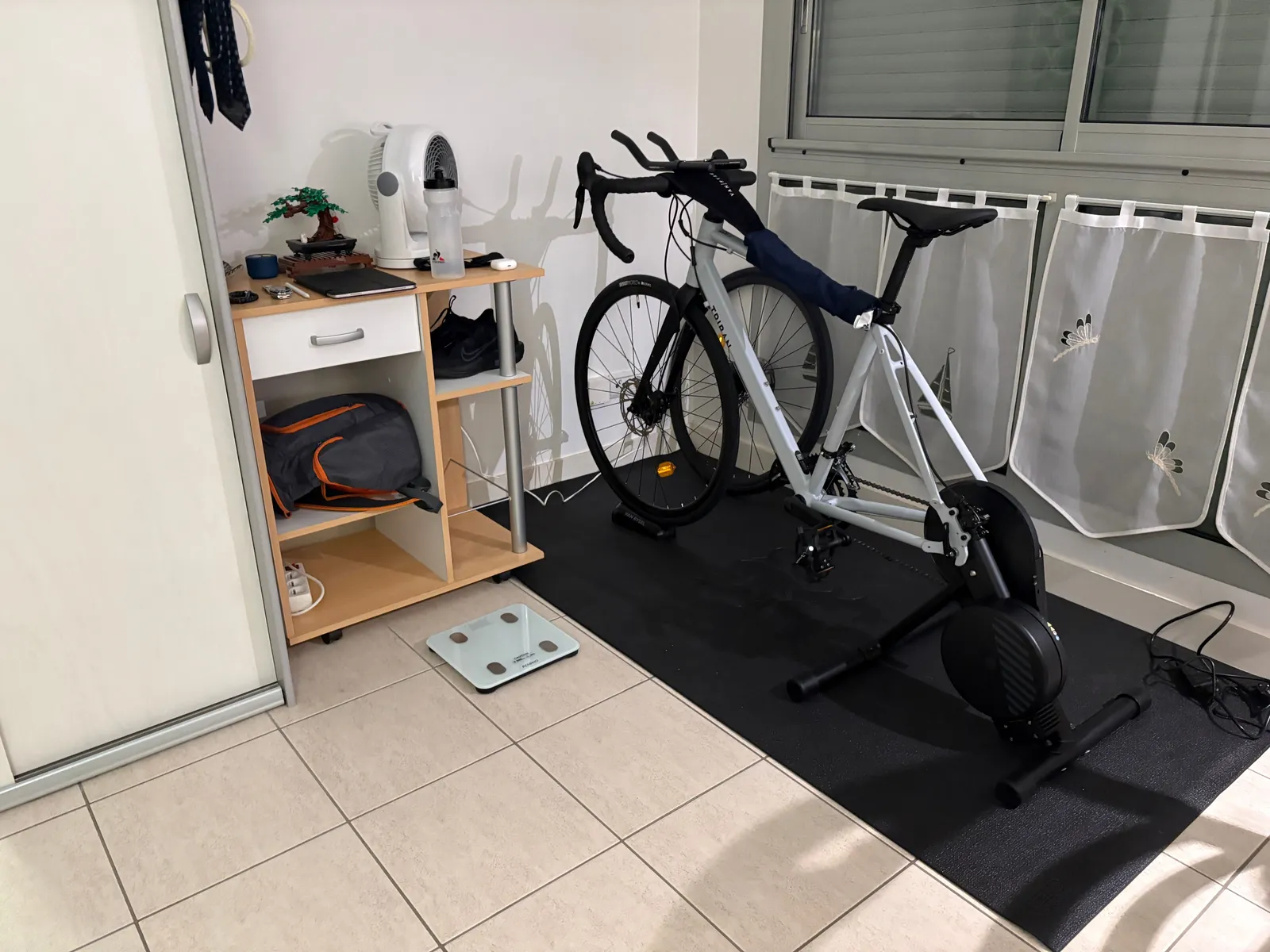An indoor smart trainer with a road bike mounted on it, set up in a spare room next to a fan and a small shelf