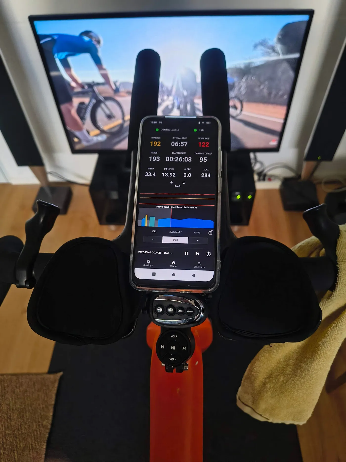 A phone on the handlebars of an indoor trainer showing an IntervalCoach Zone 2 endurance workout in the Auuki app